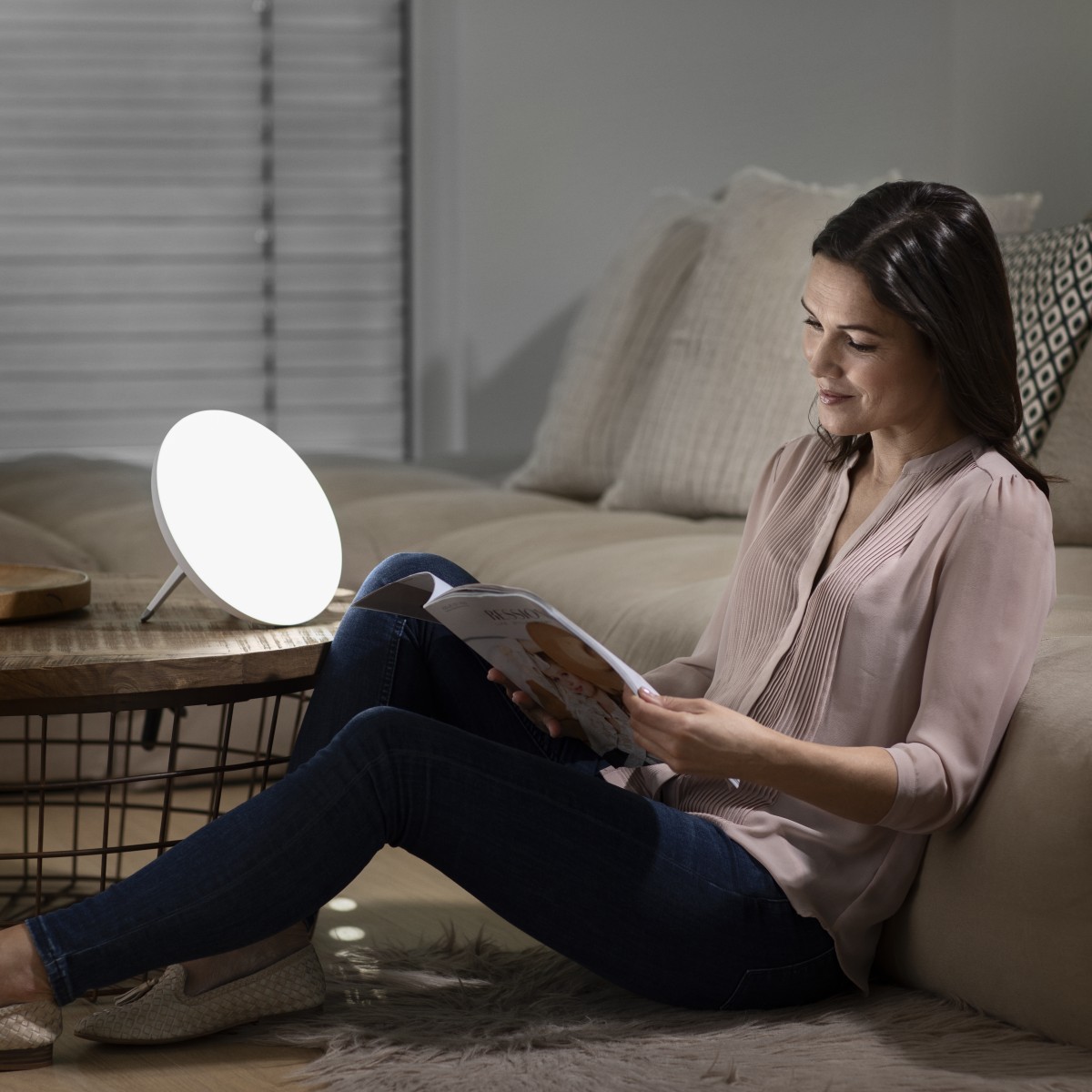 Eine Frau sitzt entspannt auf dem Boden und liest eine Zeitschrift, während eine Tageslichtlampe neben ihr leuchtet.